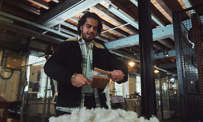 A man stands over a pile of washed woollen fleece. He is holding two wooden paddles with wire teeth, rather like oversized hairbrushes. By scraping a